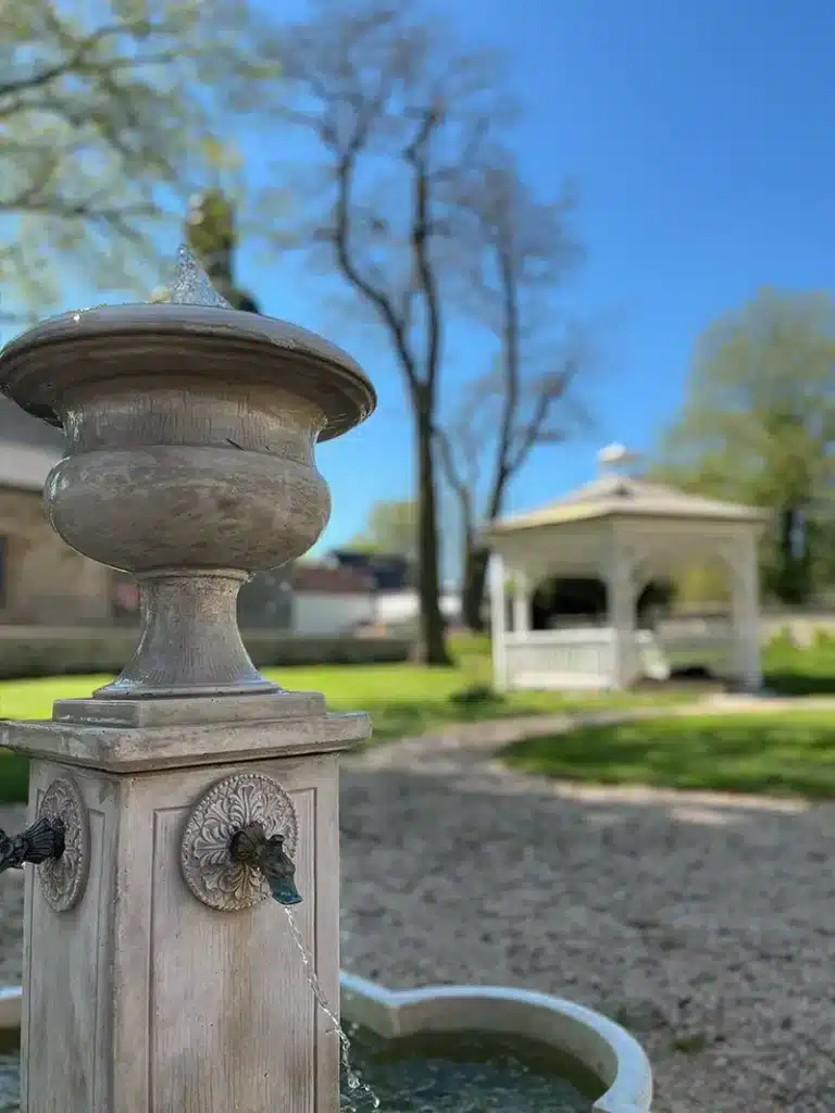 Ein steinerner Brunnen mit Wasserspeiern steht in einer Kiesfläche, perfekt für Veranstaltungen im Kyffhäuserkreis. Ein weißer Pavillon, grünes Gras, Bäume und ein strahlend blauer Himmel schaffen eine einladende Atmosphäre für den Veranstaltungsort Thüringen.