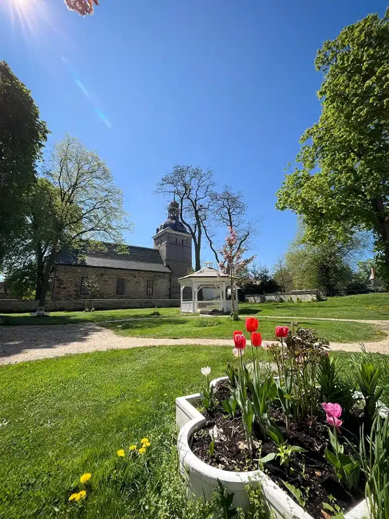 Ein sonniger Tag im Hochzeitspark Thüringen mit blühenden roten und rosafarbenen Tulpen in einem runden Pflanzgefäß, grünem Gras, Bäumen und einem steinernen Gebäude mit Kuppel und Turm unter einem klaren blauen Himmel - eine ideale Eventlocation in Thüringen.