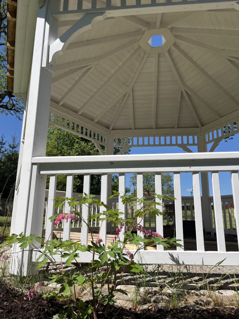 Ein weißer Holzpavillon mit einem sechseckigen Dach steht in einem Garten im Hochzeitspark Thüringen. Davor blühen rosa Herzblumen, während das Sonnenlicht Schatten auf den Pavillon und den Boden wirft. Bäume und ein blauer Himmel vervollständigen diese charmante Eventlocation in Thüringen.
