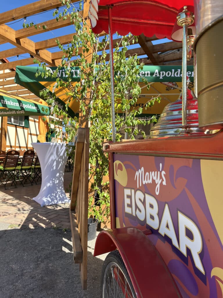 Ein bunter Eiswagen mit der Aufschrift "Mary's EISBAR" erhellt die Eventgastronomie vor der Außenbestuhlung mit hölzernen Pergolen, grünen Schirmen, Tischen und Stühlen - perfekt für jede hochzeitslocation Thüringen an einem sonnigen Tag