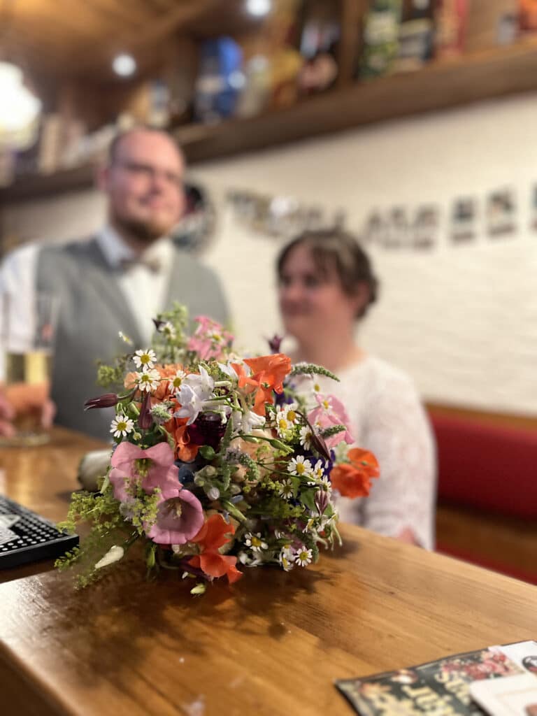 Ein farbenfroher Wildblumenstrauß liegt auf einer hölzernen Stange im Fokus, während ein lächelndes Paar in Hochzeitskleidung unscharf im Hintergrund zu sehen ist, um die Freude an einer Feier in einer Hochzeitslocation in Thüringen einzufangen