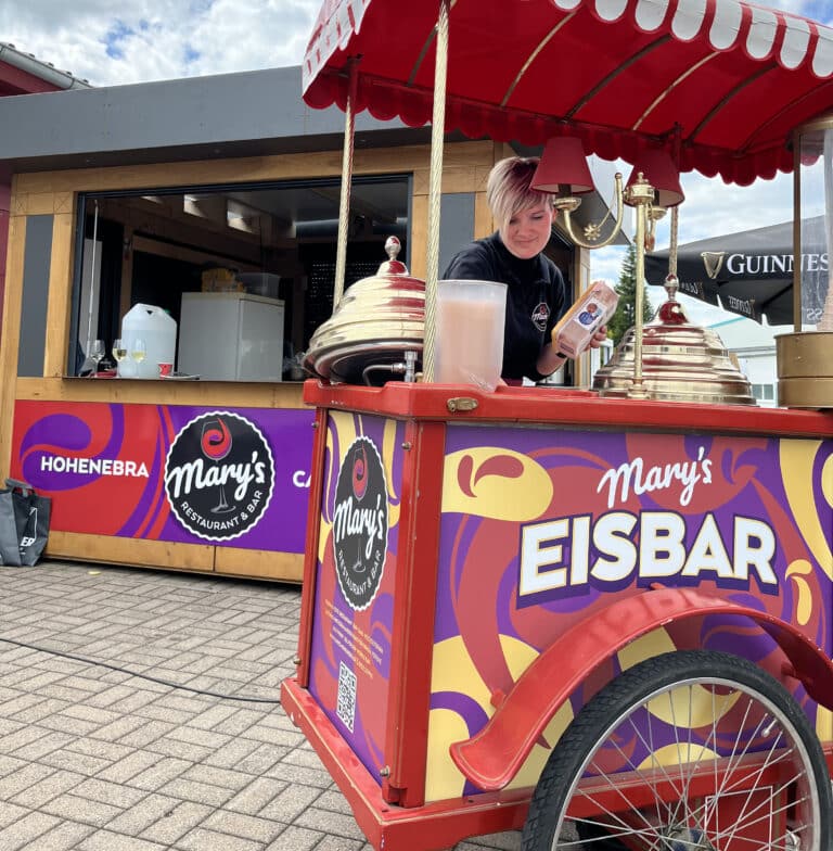 Eine Frau serviert Eis aus einem bunten Mary's EISBAR-Wagen vor einem Imbissstand mit "HOHEHEBRA"- und "Mary's"-Schildern - perfekt für Veranstaltungen oder als Hochzeitslocation Thüringen, auf einem gepflasterten Außengelände unter einem teilweise bewölkten Himmel