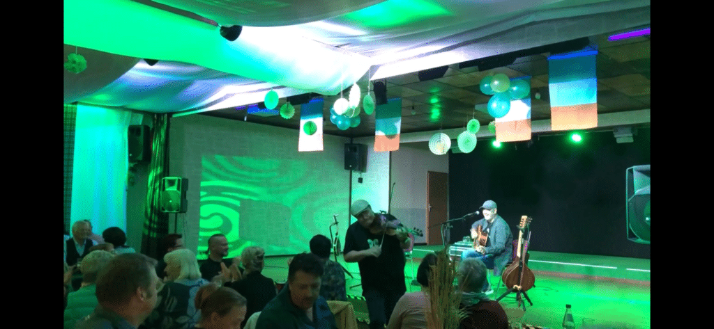 Ein Musiker spielt Gitarre und singt auf der Bühne, während ein anderer inmitten der sitzenden Zuschauer in dieser einzigartigen Eventlocation in Thüringen Geige spielt. Der Raum leuchtet mit grünen Lichtern, hängenden Luftballons und irischen Flaggen, während die Leute zusehen und sich unterhalten.