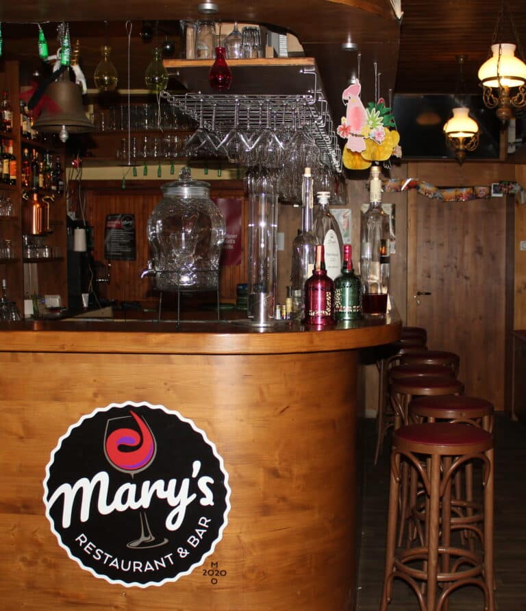 Eine Holzbar mit Hockern, Flaschen, Gläsern und einem großen Wasserspender. Auf dem Schild steht "Mary's Restaurant & Bar". Diese gemütliche Bar mit hängenden Lichtern und festlicher Dekoration ist eine charmante Hochzeitslocation in Thüringen für jede Feier