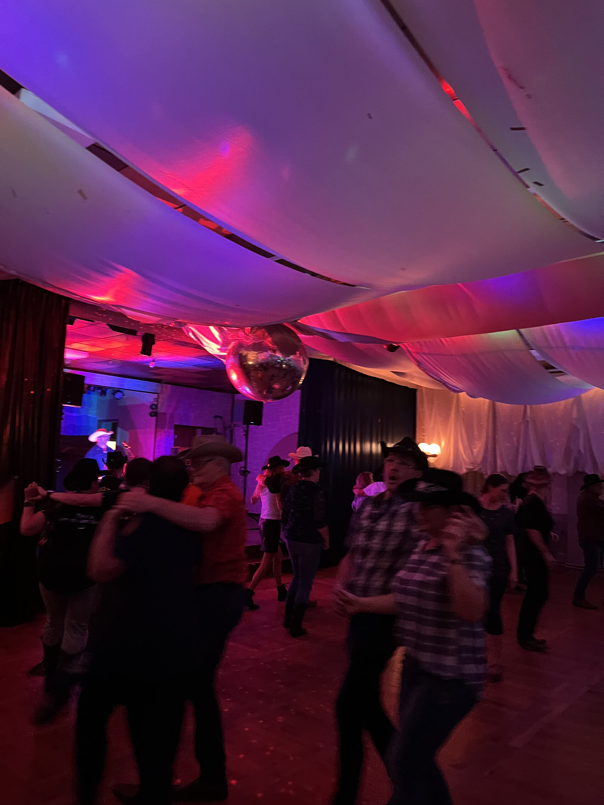 Die Leute tanzen in einem schwach beleuchteten Raum mit einer Discokugel, bunten Lichtern und drapierten Deckenstoffen. Die meisten tragen Cowboyhüte und legere Kleidung, was zu der lebhaften, an das Westernthema angelehnten Atmosphäre der Line Dance Party beiträgt.