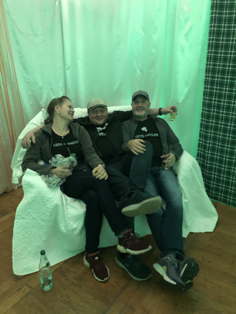 Drei Freunde sitzen eng beieinander auf einer weißen Couch und lächeln und lachen im grünen Licht des hochzeitsparks Thüringen. Entspannt in legerer Kleidung genießen sie die fröhliche Stimmung dieser Top-Hochzeitslocation in Thüringen, während eine Flasche Wasser auf dem Boden steht.