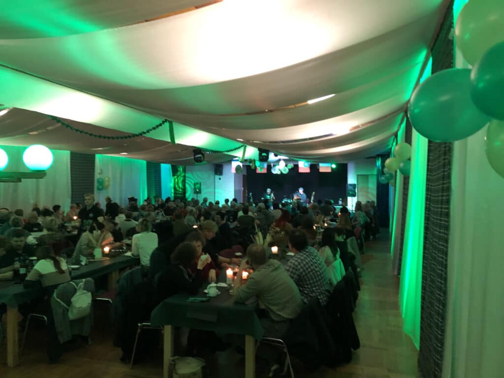 Eine große Gruppe sitzt an geschmückten Tischen bei grüner Beleuchtung in einem geräumigen Saal im Hochzeitspark Thüringen und genießt Essen und Gespräche, während eine Band spielt. Luftballons und drapierte Stoffe schaffen eine festliche Event-Gastronomie-Atmosphäre.