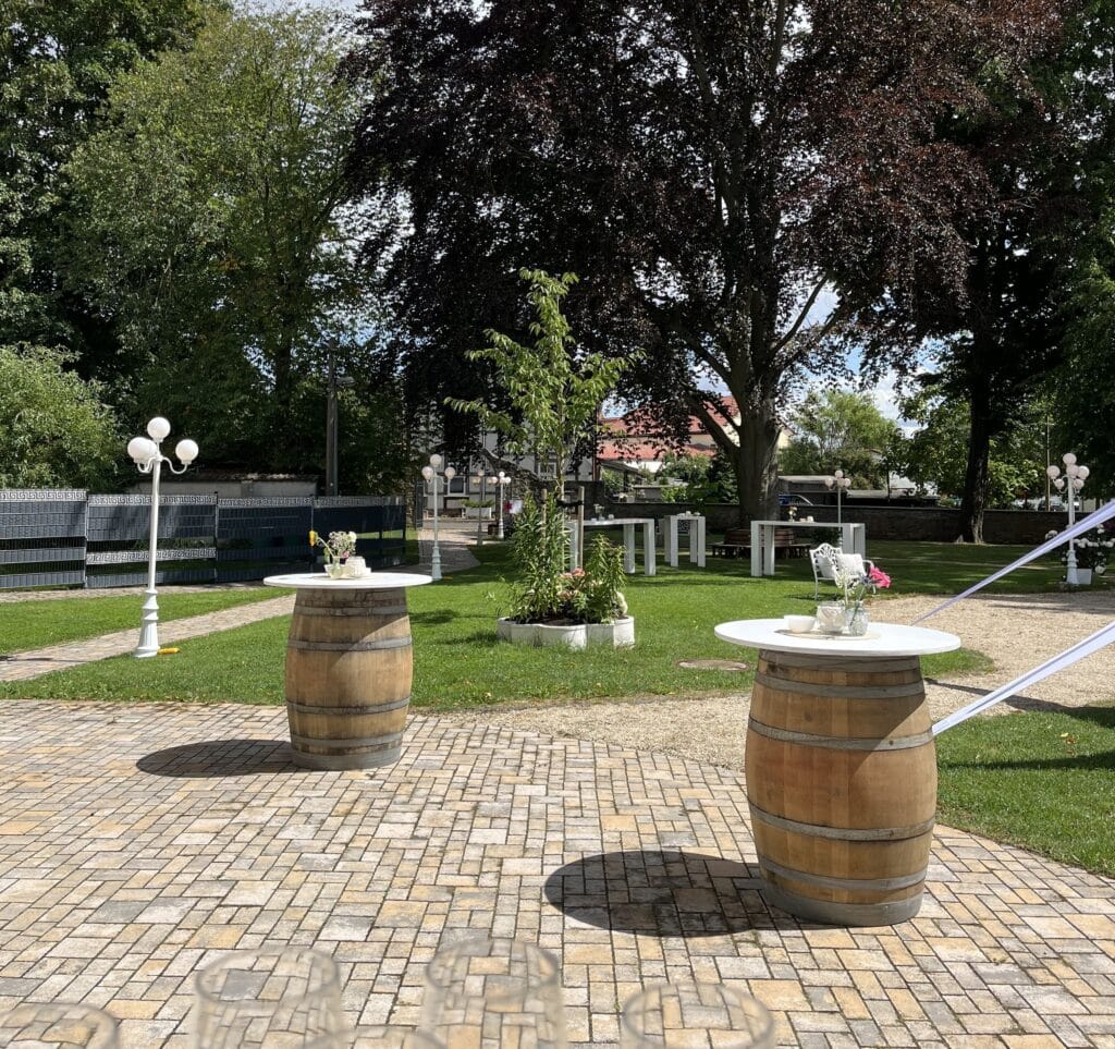 Sonnige Außenterrasse mit zwei hohen, blumengeschmückten Tischen, perfekt für eine Trauung im Freien. Umgeben von Gras, Bäumen, weißen Bänken und dekorativen Lichtern, mit einem Weg und Zaun im Hintergrund.