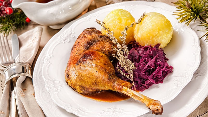 Ein festlicher Teller mit gebratener Entenkeule, Kartoffelklößen und geschmortem Rotkohl wird auf edlem weißen Porzellan serviert - perfekt für besondere Anlässe in einer Eventlocation in Thüringen mit weihnachtlicher Dekoration und elegantem Silberbesteck.