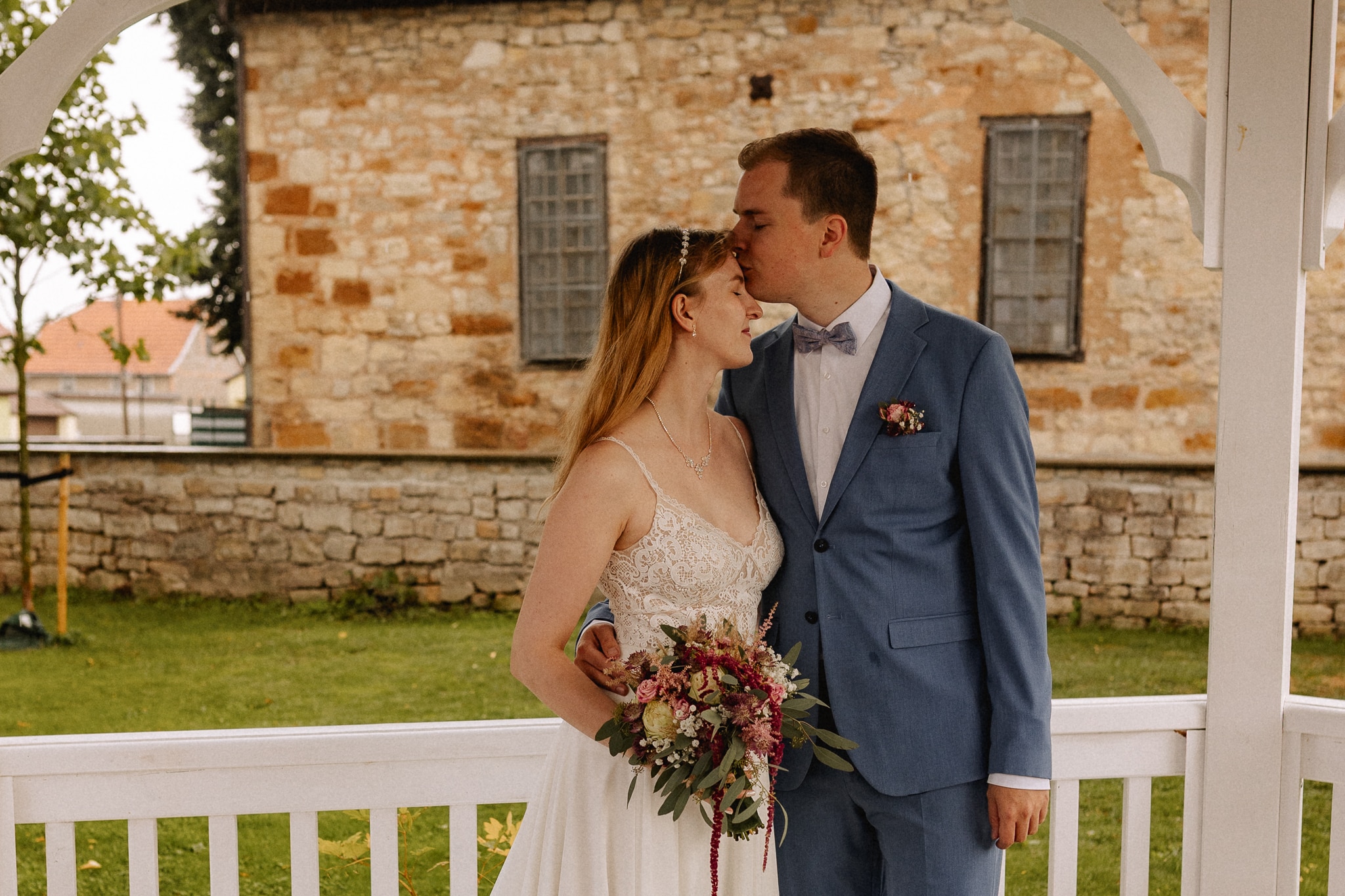Ein Bräutigam in einem blauen Anzug küsst die Stirn seiner Braut in einem weißen Kleid, die einen Blumenstrauß in der Hand hält. Sie stehen zusammen unter einem weißen Pavillon mit einem Steingebäude im Hintergrund und fangen Momente ein, die direkt aus einer fröhlichen Hochzeitsnachricht stammen.