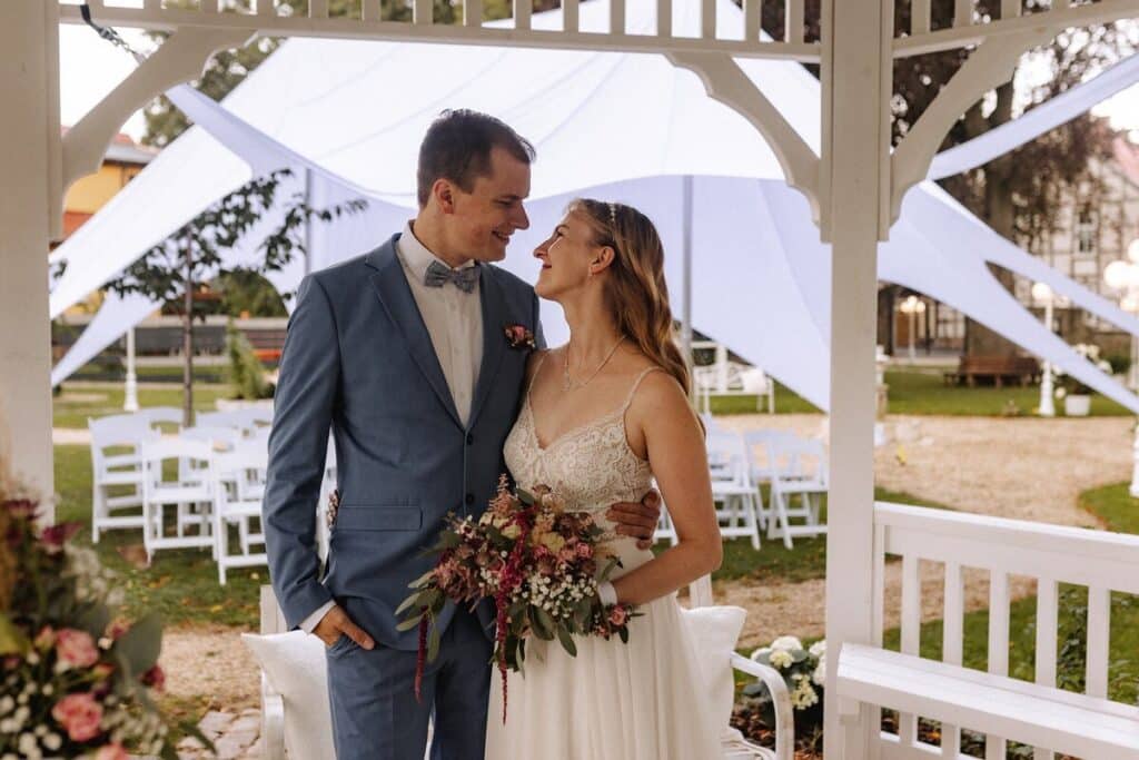 Eine Braut und ein Bräutigam stehen dicht beieinander in einem Pavillon im Freien in einer Eventlocation in Thüringen, schauen sich an und lächeln. Der Bräutigam trägt einen hellblauen Anzug, die Braut ein weißes Kleid mit einem Blumenstrauß. Weiße Stühle und ein Zelt vervollständigen die Szene.