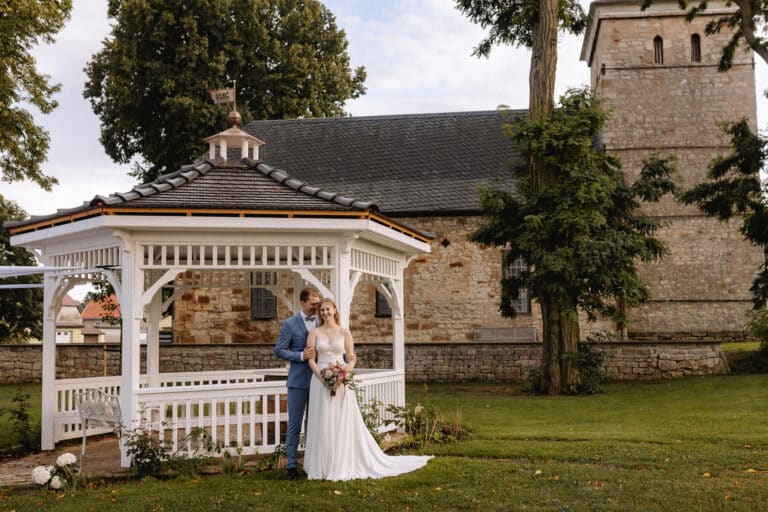 Eine Braut und ein Bräutigam stehen unter einem weißen Pavillon in einem grasbewachsenen Garten in der schönen Eventlocation Thüringen, mit einer Steinkirche und hohen Bäumen im Hintergrund. Die Braut hält einen Blumenstrauß und trägt ein weißes Kleid, der Bräutigam trägt einen blauen Anzug
