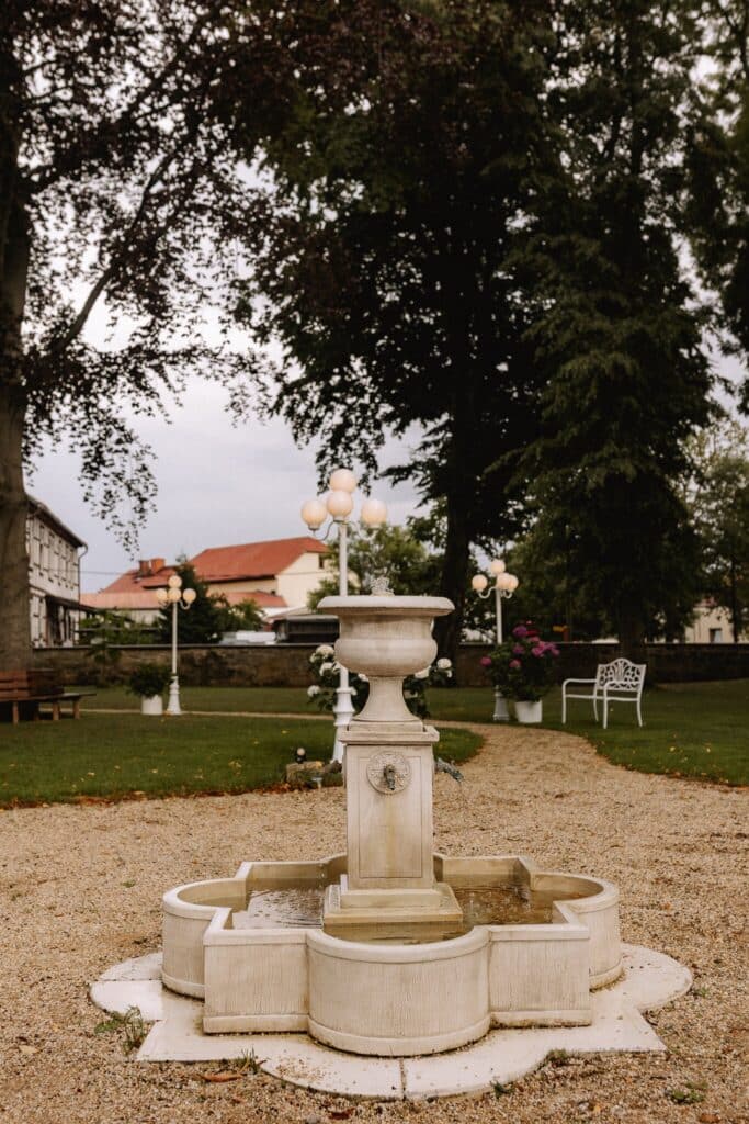 Ein weißer Steinbrunnen mit dekorativen Lichtern steht auf einem Kiesweg in einem Garten in einer Eventlocation in Thüringen, umgeben von grünem Gras, Bäumen, Bänken und einer weißen, verzierten Bank. Im Hintergrund sind Gebäude mit roten Dächern zu sehen.
