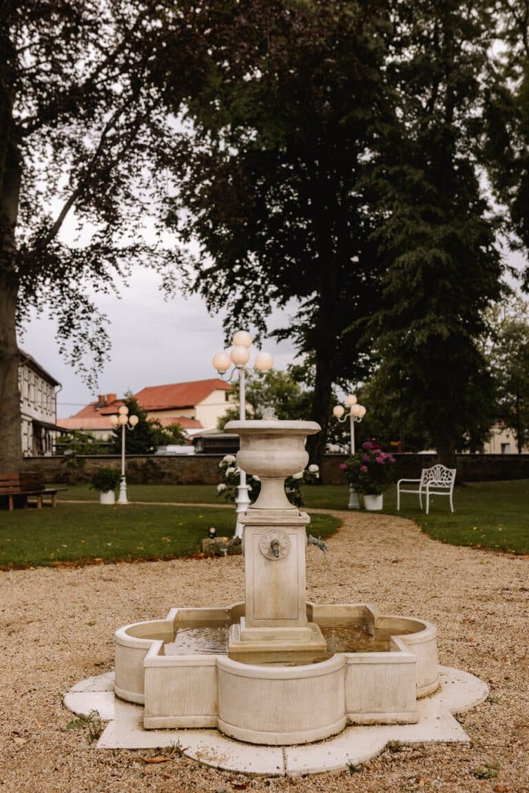 Ein weißer Steinbrunnen mit dekorativen Lichtern steht auf einem Kiesweg in einem Garten in einer Eventlocation in Thüringen, umgeben von grünem Gras, Bäumen, Bänken und einer weißen, verzierten Bank. Im Hintergrund sind Gebäude mit roten Dächern zu sehen