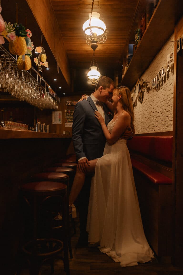 Eine Braut und ein Bräutigam teilen einen intimen Moment in einer schwach beleuchteten Bar in der Eventlocation Thüringen. Die Braut in einem weißen Kleid auf einem Barhocker und der Bräutigam in einem blauen Anzug stehen dicht beieinander, berühren sich an der Stirn und lächeln
