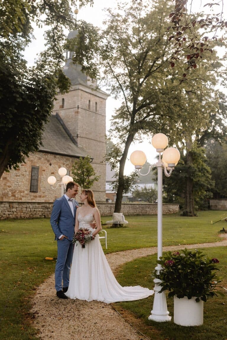 Eine Braut in einem weißen Kleid und ein Bräutigam in einem blauen Anzug stehen zusammen auf einem Gartenweg in der malerischen Eventlocation Thüringen, umgeben von Grün und Laternenpfählen, mit einem steinernen Kirchengebäude im Hintergrund