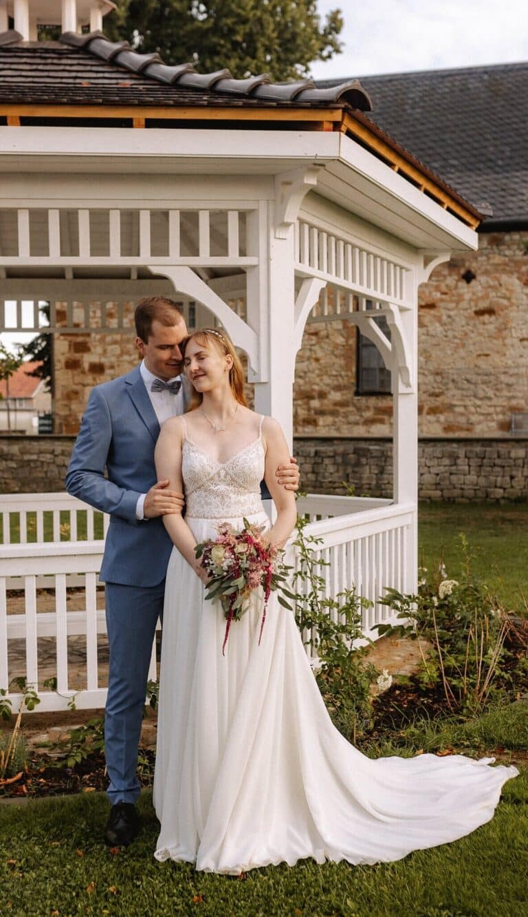 Eine Braut und ein Bräutigam stehen gemeinsam vor einem weißen Pavillon in der Eventlocation Thüringen. Die Braut in einem weißen Kleid hält einen Blumenstrauß, während der Bräutigam in einem hellblauen Anzug sie von hinten umarmt. Beide sehen zufrieden und glücklich aus