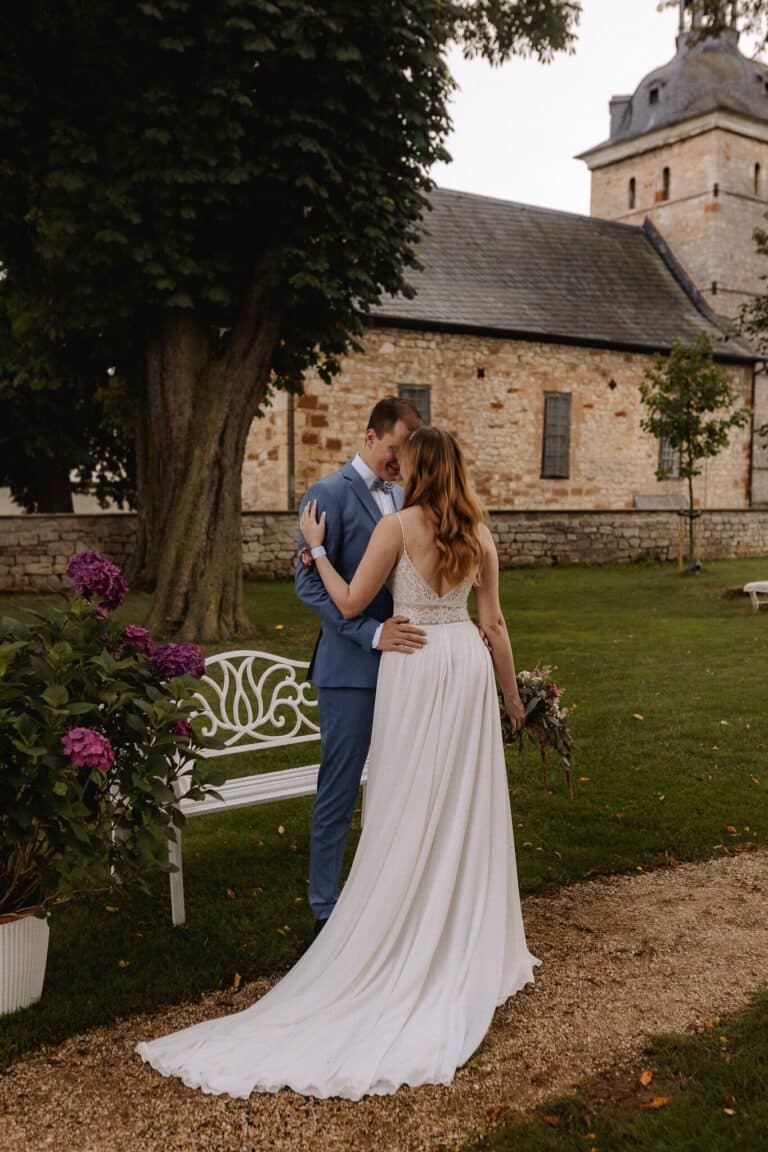 Eine Braut in einem weißen Kleid und ein Bräutigam in einem blauen Anzug umarmen sich im Freien in der Nähe einer weißen Bank in einer Eventlocation in Thüringen. Ein Steinhaus, Grünpflanzen und der Brautstrauß vervollständigen die romantische Szene