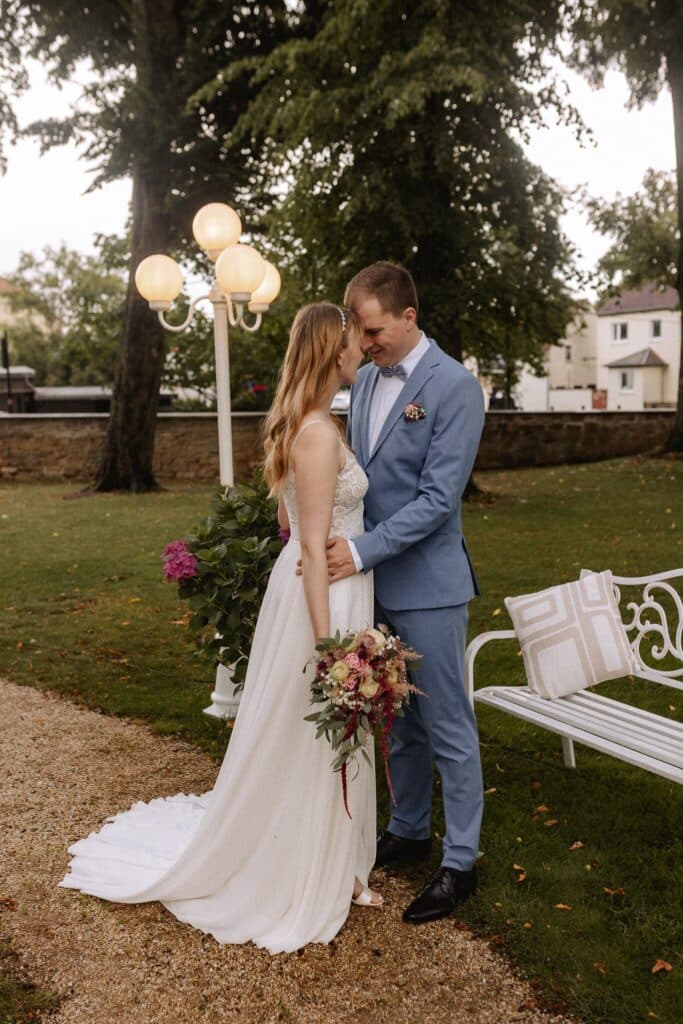 Eine Braut in einem weißen Kleid mit einem Blumenstrauß in der Hand steht eng mit einem Bräutigam in einem hellblauen Anzug in einem Garten der Eventlocation Thüringen, in der Nähe einer weißen Bank und eines Laternenpfahls, umgeben von Bäumen und charmanten Häusern.