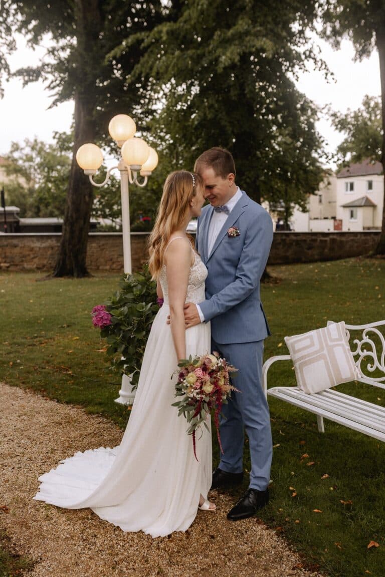 Eine Braut in einem weißen Kleid mit einem Blumenstrauß in der Hand steht eng mit einem Bräutigam in einem hellblauen Anzug in einem Garten der Eventlocation Thüringen, in der Nähe einer weißen Bank und eines Laternenpfahls, umgeben von Bäumen und charmanten Häusern