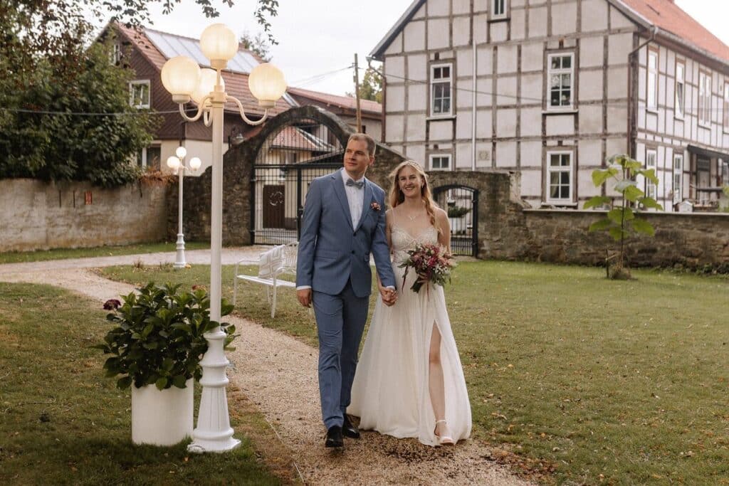 Eine Braut und ein Bräutigam gehen Arm in Arm einen Kiesweg in einem Garten in einer Eventlocation in Thüringen entlang, gekleidet in ein weißes Kleid und einen blauen Anzug, mit einem rustikalen Gebäude und Straßenlaternen im Hintergrund. Die Braut hält einen Blumenstrauß in der Hand und lächelt.