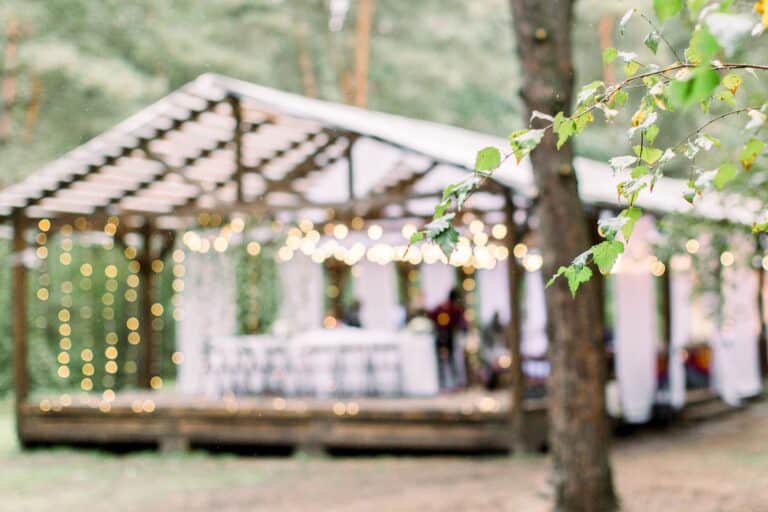 Ein verschwommener, mit Lichterketten und weißen Vorhängen geschmückter Festzelt steht zwischen Bäumen, im Vordergrund ein Ast mit grünen Blättern - eine bezaubernde Eventlocation Thüringen für Hochzeiten im Zelt oder heiraten im eigenen Garten.