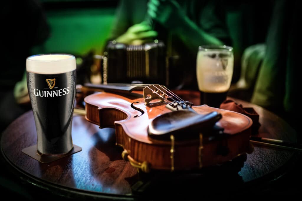 Ein Pint Guinness steht auf einem Holztisch neben einer Geige und einem Bogen, ein weiteres Pint und ein verschwommener Musiker, der im Hintergrund Ziehharmonika spielt, beschwören die gemütliche Kneipenstimmung herauf, die perfekt für eine Feier der Freien Trauung Thüringen ist.