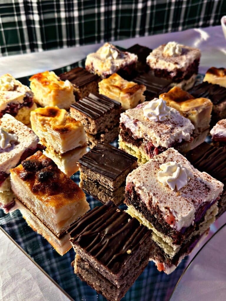 Auf einer verspiegelten Fläche mit kariertem Hintergrund steht ein Tablett mit verschiedenen Gebäckstücken, darunter mit Schokolade überzogene Quadrate, Schichtkuchen mit Sahne und Früchten sowie Riegel mit Vanillecreme