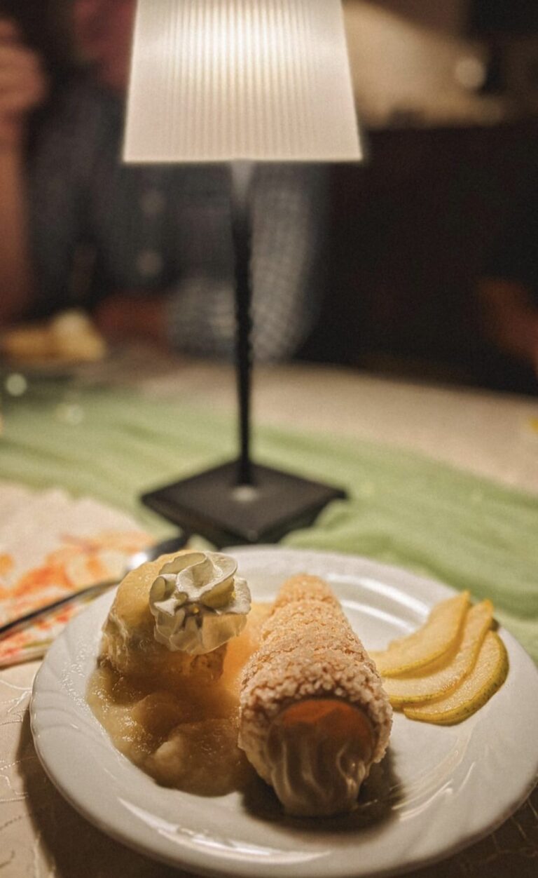 Ein Teller mit einem mit Sahne gefüllten Gebäck, Birnenscheiben und einer Kugel Eis mit Schlagsahne steht unter einer kleinen Tischlampe auf einem dekorierten Tisch. Eine Person sitzt unscharf im Hintergrund