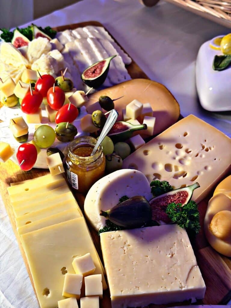 Ein Holzbrett mit verschiedenen Käsesorten, Weintrauben, Feigen, Kirschtomaten am Spieß, Oliven, Petersilie und einem Glas Marmelade mit einem Löffel, arrangiert als Vorspeisenteller