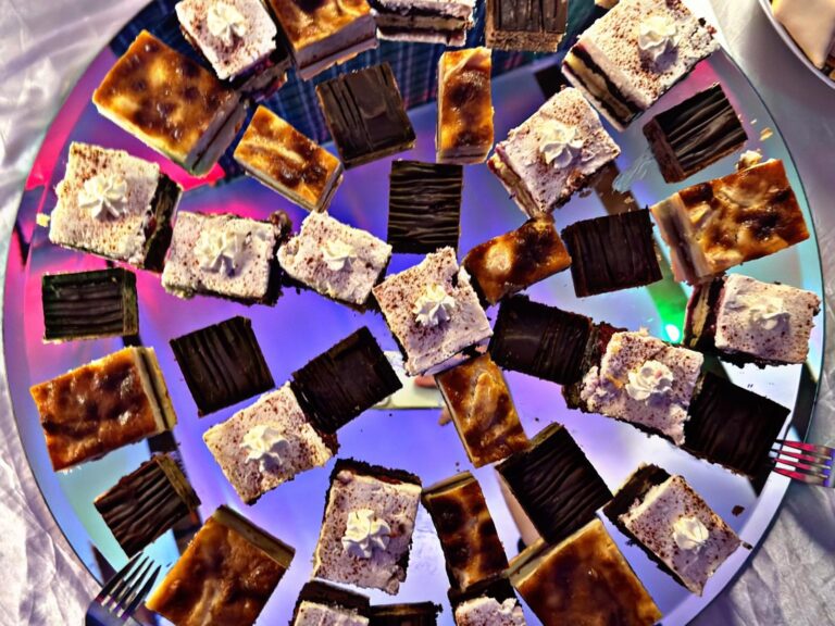 Eine Auswahl an rechteckigen Kuchen und Gebäck mit verschiedenen Belägen, die in einem kreisförmigen Muster auf einem reflektierenden, schillernden Tablett angeordnet sind, wobei an den Seiten kleine Gabeln zu sehen sind