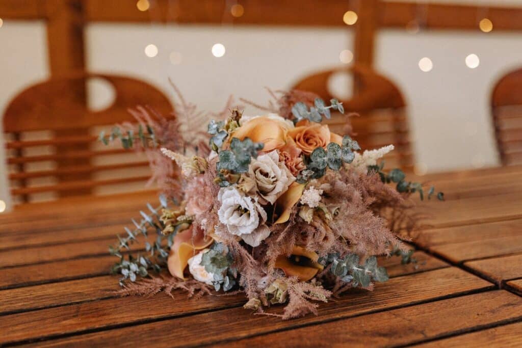 Ein rustikaler Strauß aus getrockneten und frischen Blumen, darunter Rosen und Eukalyptus, steht auf einem Holztisch. Im Hintergrund sind verschwommene Holzstühle und Lichterketten zu sehen, die eine warme, gemütliche Atmosphäre schaffen.