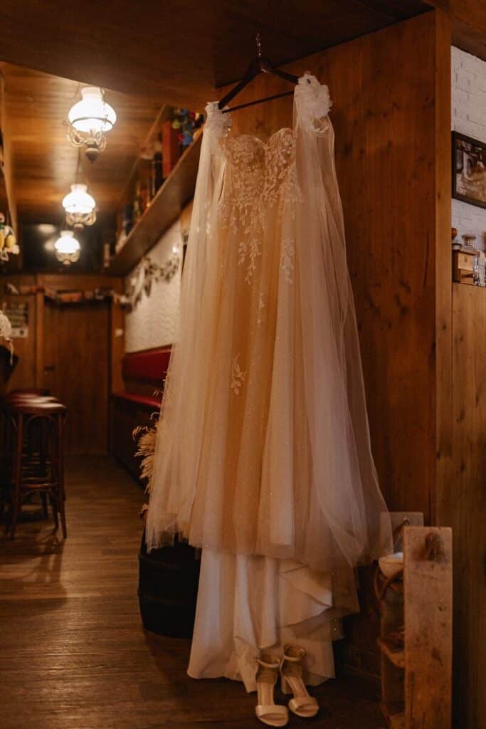 Ein langes, elegantes Hochzeitskleid mit Spitzendetails hängt an einer Holzwand in einem gemütlichen, warm beleuchteten Raum. Ein Paar weißer hochhackiger Schuhe steht auf dem Boden unter dem Kleid.