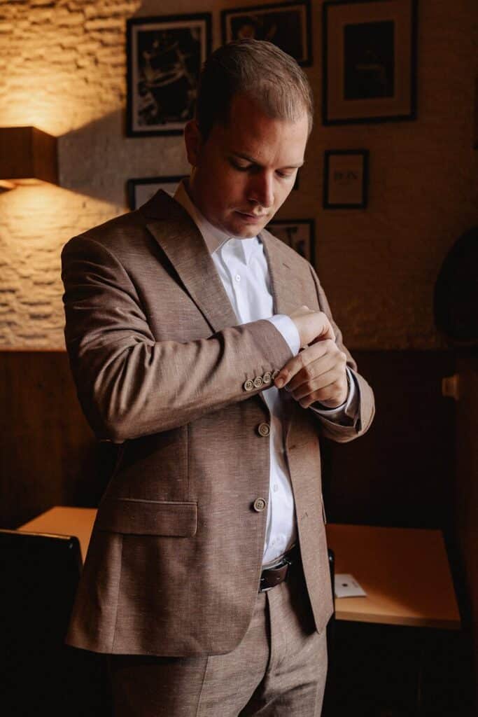 Ein Mann in einem braunen Anzug steht in einem Raum, blickt nach unten und rückt seine Hemdmanschette zurecht. Er befindet sich in einem warm beleuchteten Raum mit gerahmten Bildern an der Wand hinter ihm.