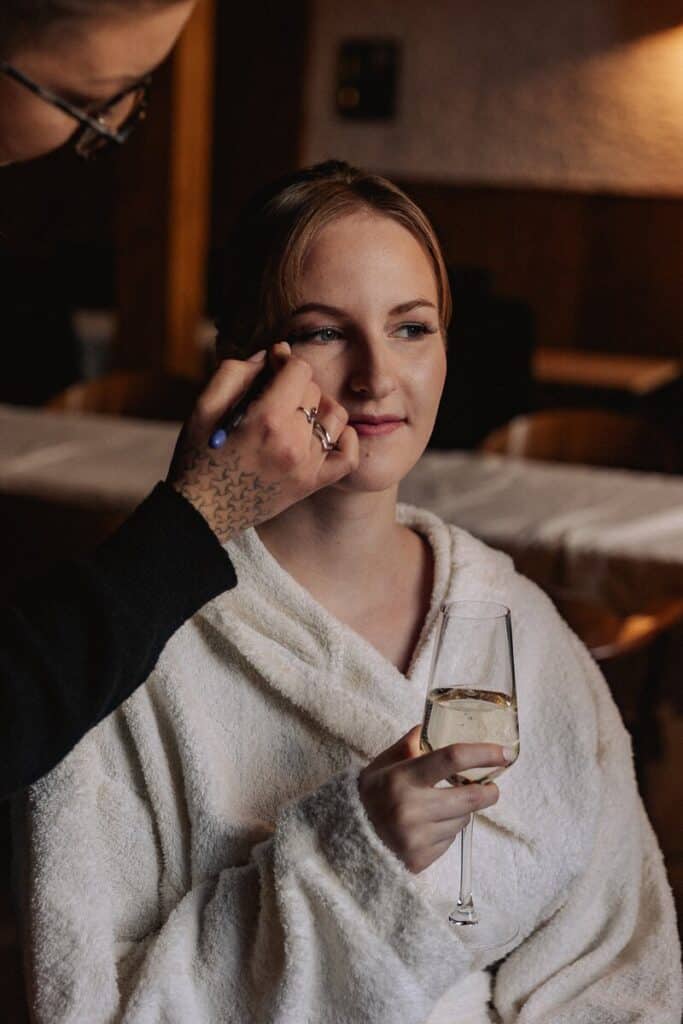 Eine Frau in einem weißen Bademantel hält ein Glas Weißwein in der Hand, während eine andere Person in einem sanft beleuchteten Raum Make-up auf ihr Gesicht aufträgt.