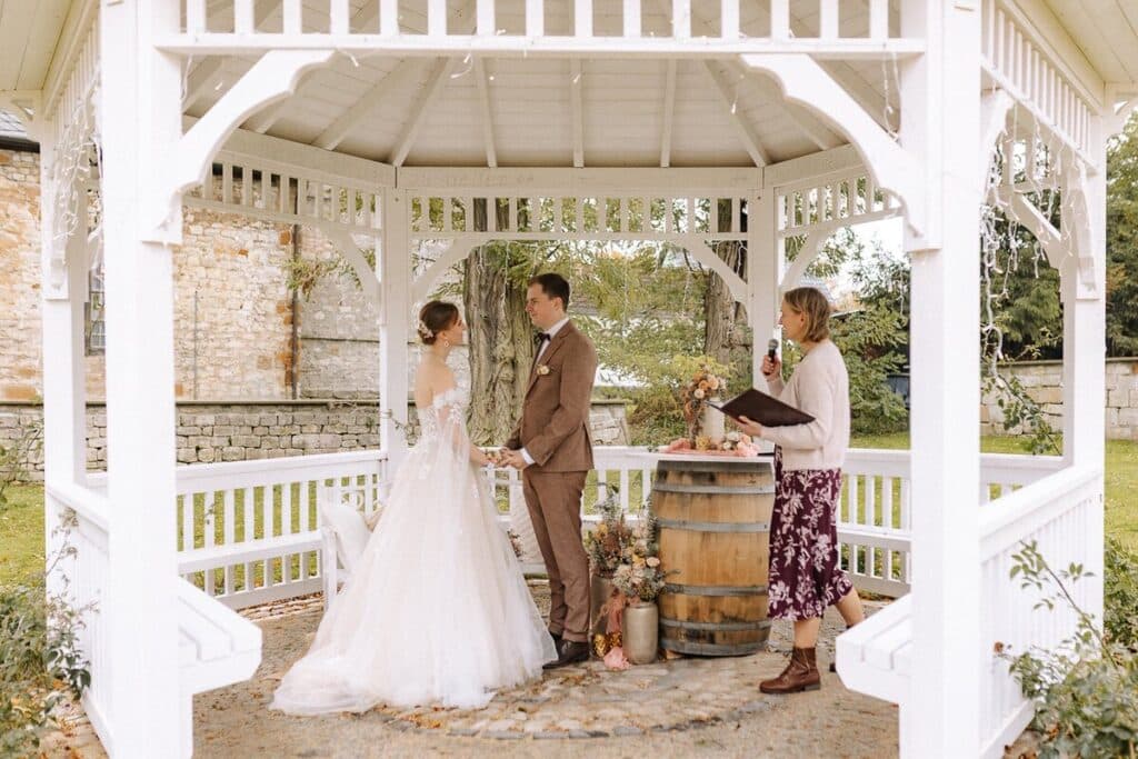 Eine Braut und ein Bräutigam stehen sich während ihrer Hochzeitszeremonie im Freien in einem weißen Pavillon gegenüber, während ein Geistlicher aus einem Buch liest. Neben den beiden steht ein Holzfass mit Blumen.