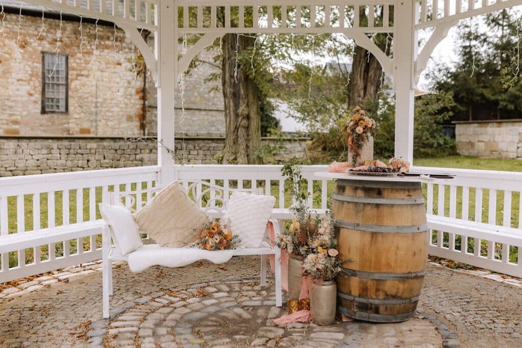 Ein gemütlicher weißer Pavillon mit einer gepolsterten Bank, Blumenarrangements und einem mit Blumen geschmückten Holztisch auf einem Kopfsteinpflaster in einem Garten mit Steinhäusern im Hintergrund.