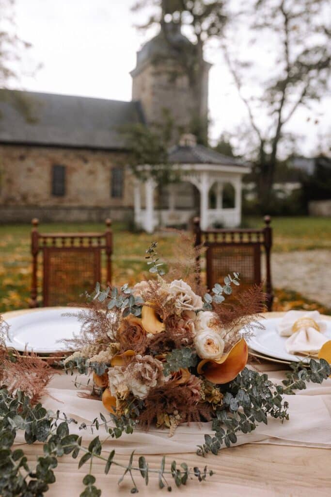 Ein rustikaler Tisch im Freien mit einem Strauß aus beigen, gelben und orangefarbenen Blumen, Grünzeug und Pampasgras. Zwei verzierte Stühle und ein Pavillon stehen vor einem Steingebäude, umgeben von Herbstlaub.