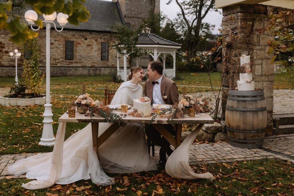 Eine Braut und ein Bräutigam sitzen an einem geschmückten Tisch im Freien und teilen einen freudigen Moment. Der Tisch ist mit Blumen und Desserts geschmückt, und in der Nähe steht eine mehrstöckige Hochzeitstorte auf einem Fass. Herbstlaub und rustikale Gebäude umgeben die beiden.