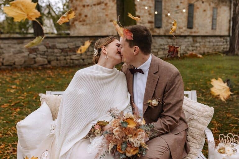 Eine Braut und ein Bräutigam sitzen auf einer weißen Bank im Freien und küssen sich, während um sie herum Herbstblätter fallen. Die Braut hält einen Blumenstrauß und trägt einen weißen Schal, der Bräutigam trägt einen braunen Anzug und eine Fliege. Im Hintergrund ist ein Steingebäude zu sehen
