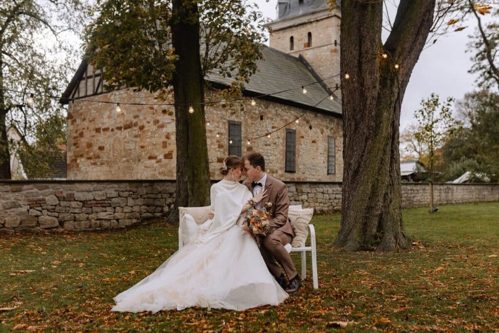 Eine Braut und ein Bräutigam sitzen eng beieinander auf einer Bank unter Lichterketten in einer grünen Umgebung, mit einer alten Steinkirche im Hintergrund. Die Braut hält einen Blumenstrauß in der Hand und trägt ein weißes Kleid; der Bräutigam trägt einen braunen Anzug.