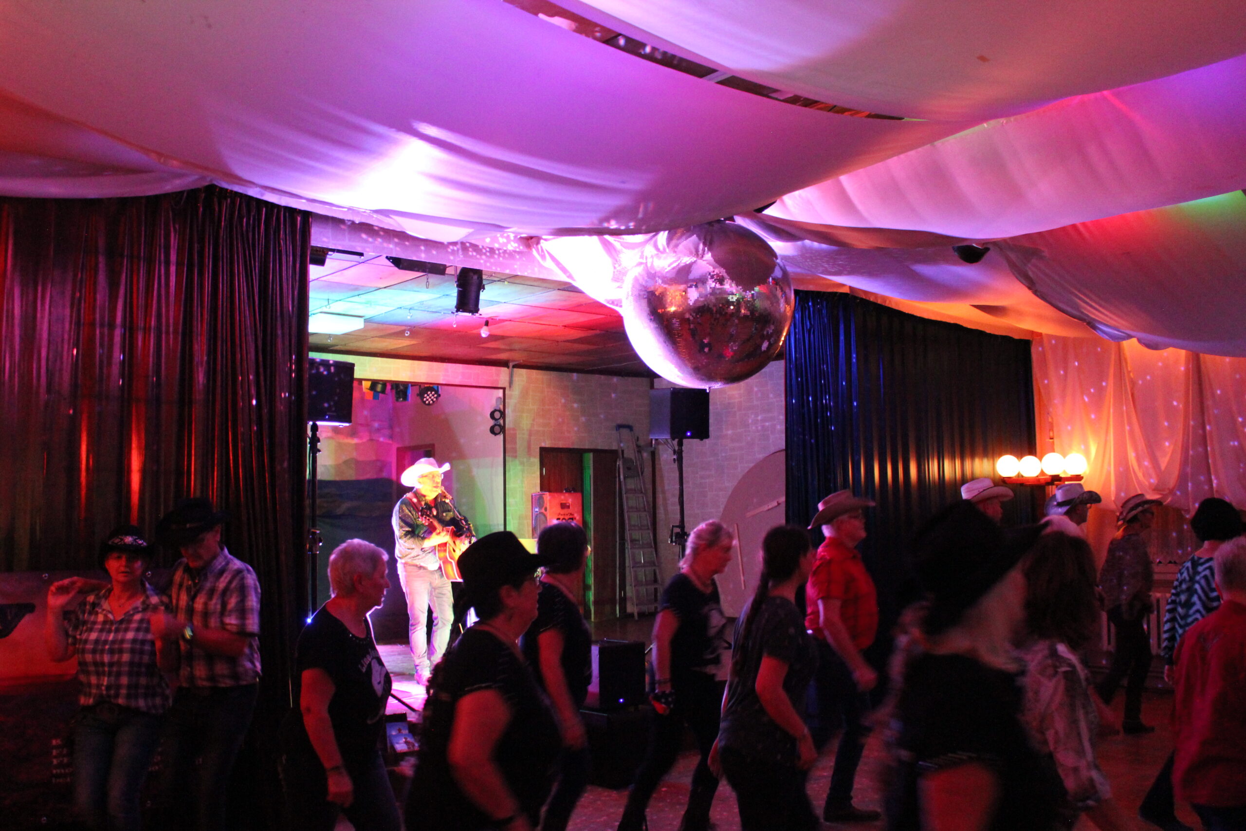 Menschen mit Cowboyhüten tanzen in einer schummrig beleuchteten Eventlocation in Thüringen mit bunten Lichtern, einer Discokugel und drapiertem weißen Stoff an der Decke. Im Hintergrund tritt ein Musiker mit Cowboyhut auf der Bühne auf.