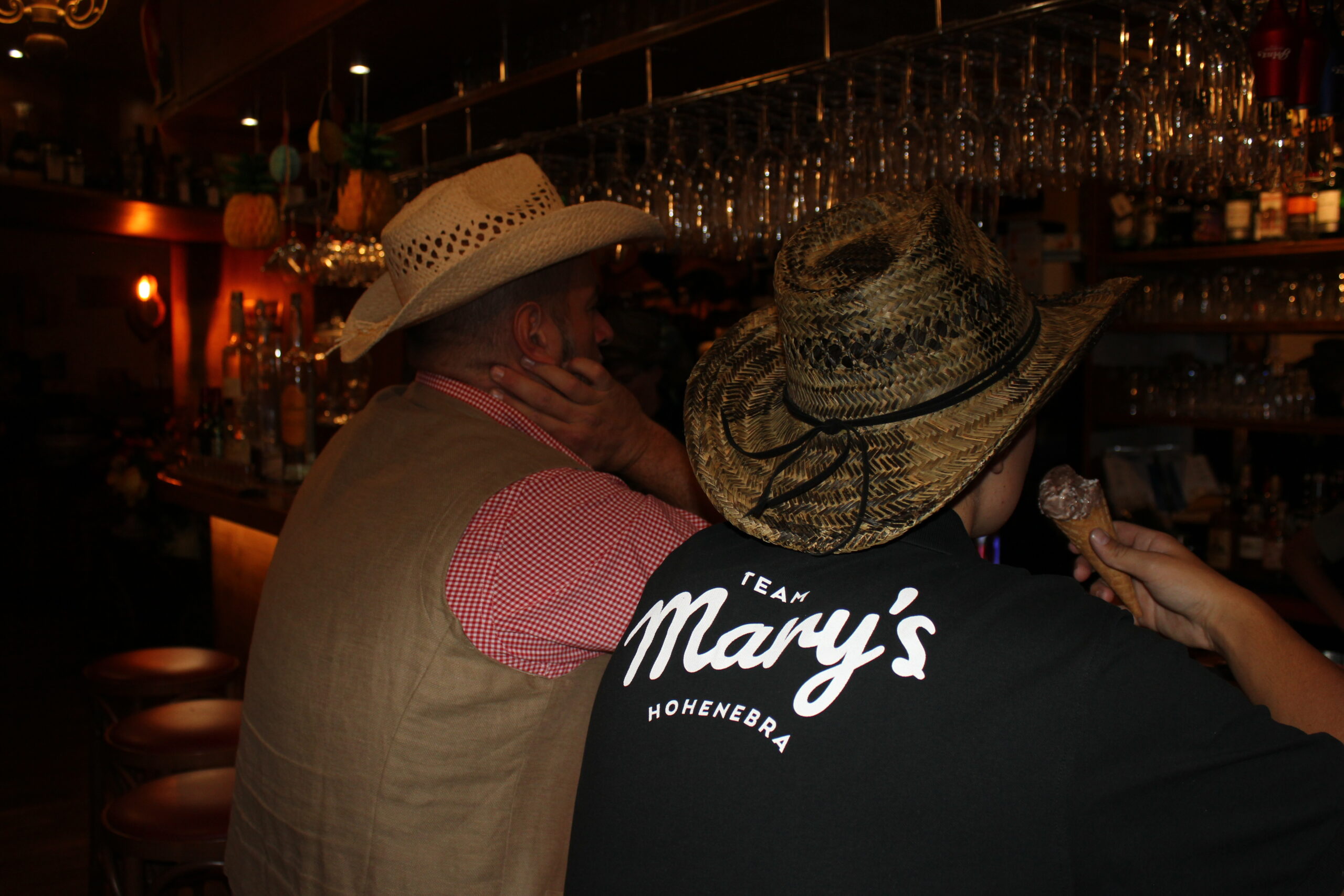 Zwei Personen mit Cowboyhüten sitzen an einer schummrigen Bar. Einer trägt eine beige Weste und ein kariertes Hemd, der andere ein schwarzes Hemd mit der Aufschrift "Team Mary's HOHENEBRA" auf dem Rücken. Gläser hängen über dem Kopf, und eine Person hält eine Eistüte in der Hand.