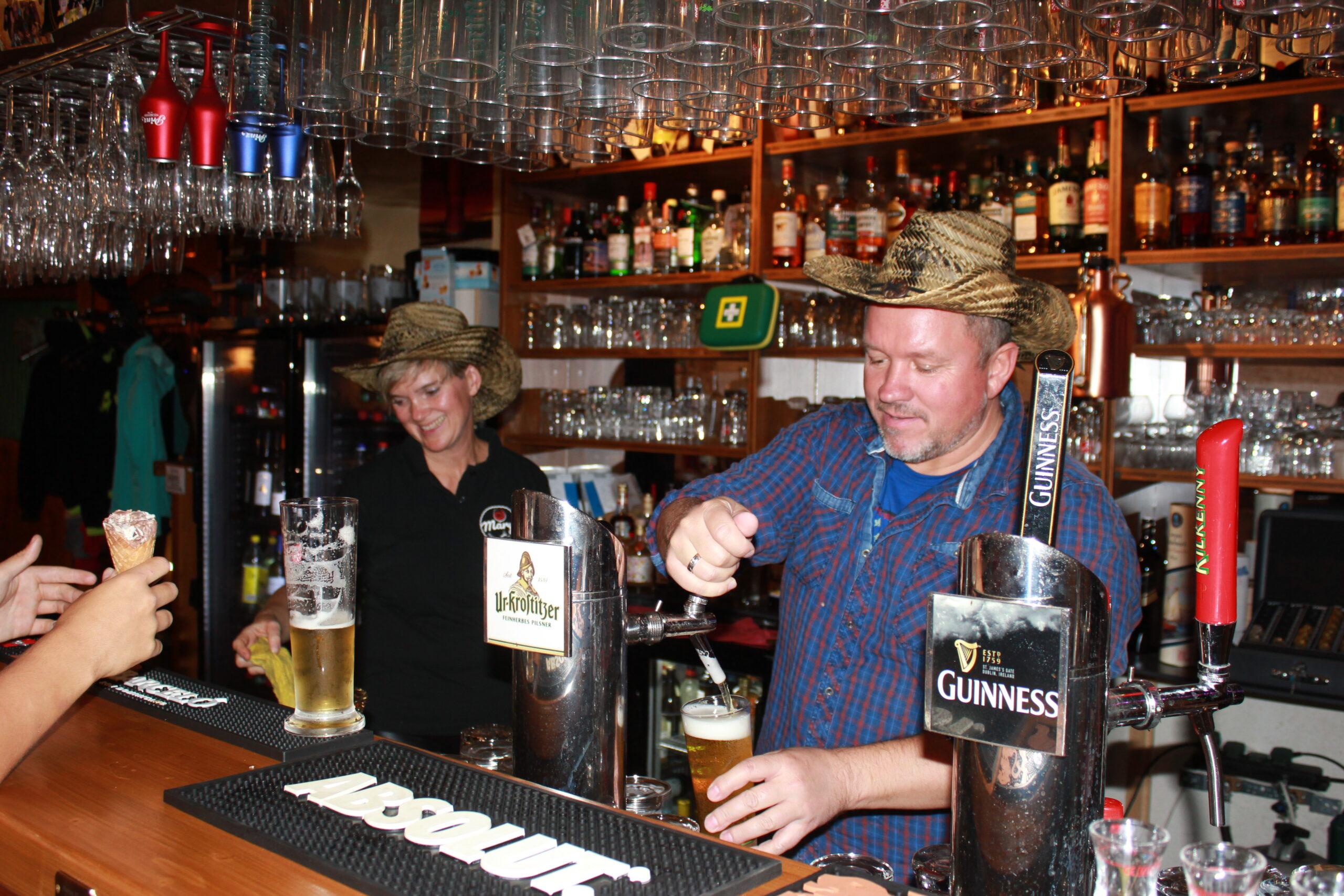 Zwei Barkeeper mit Strohhüten arbeiten hinter einer Bar in einer Eventlocation in Thüringen und schenken Getränke aus Zapfhähnen ein. Der eine schenkt lächelnd Bier ein, der andere bedient einen Kunden mit einer Waffeltüte. Regale mit Gläsern und Flaschen säumen den Hintergrund.