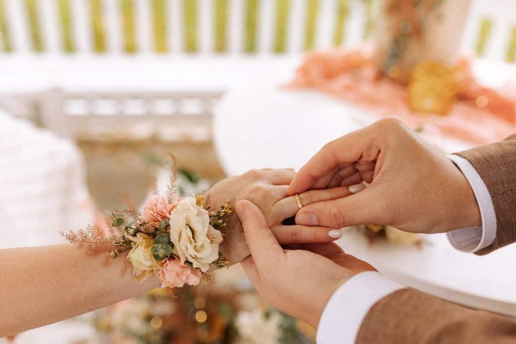 Eine Person in einem Anzug steckt einer anderen Person, die ein Handgelenkkorsett aus rosa und weißen Blumen trägt, vorsichtig einen Ring an den Finger. Der Hintergrund ist sanft verschwommen und suggeriert eine romantische Umgebung oder eine Hochzeit.