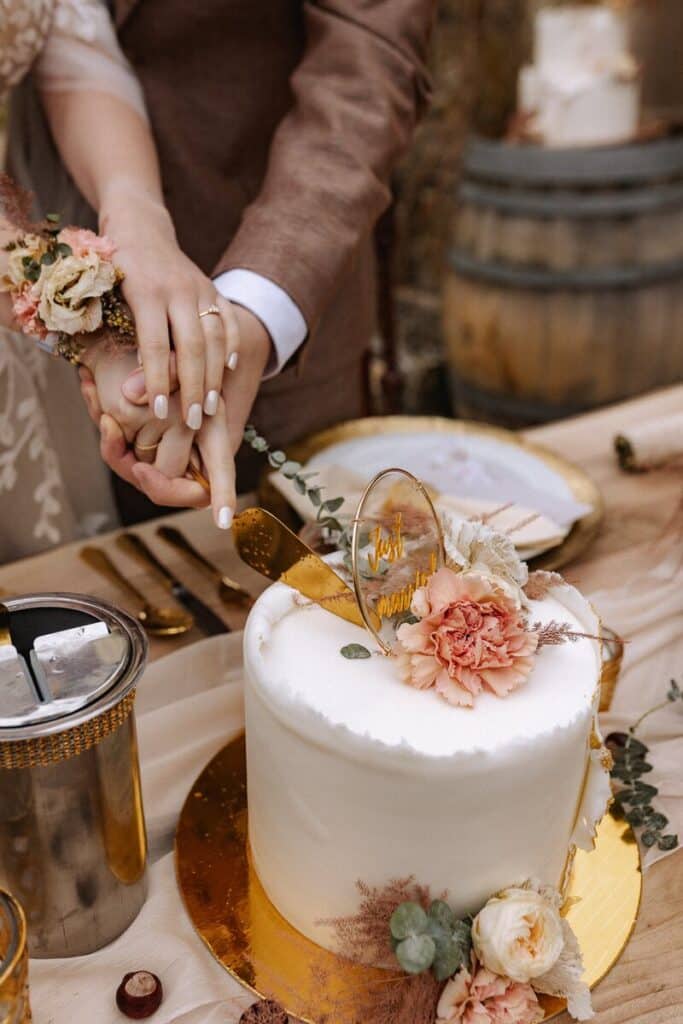Ein Paar in Hochzeitskleidung schneidet eine weiße Torte an, die mit Blumen und einem goldenen "Just Married"-Schriftzug verziert ist. Ihre Hände, die beide Ringe tragen, halten das Messer zusammen. Der Tisch ist mit Tellern, Besteck und rustikaler Dekoration gedeckt.