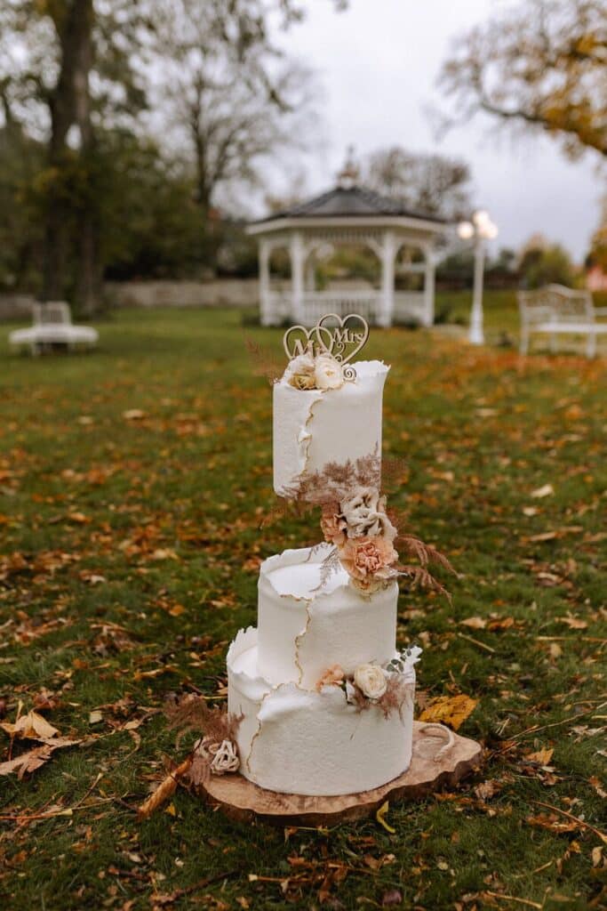 Eine dreistöckige weiße Hochzeitstorte mit goldenen Akzenten und Trockenblumen steht auf einer Wiese in einem Park, in dem Herbstblätter verstreut sind und ein weißer Pavillon im Hintergrund zu sehen ist.