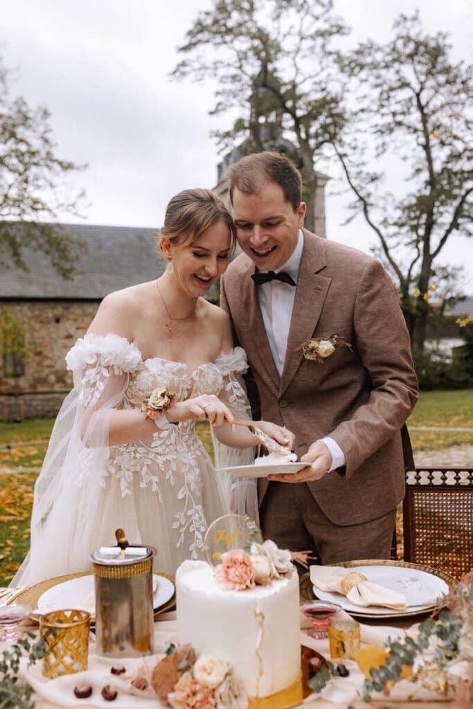 Eine lächelnde Braut und ein lächelnder Bräutigam stehen im Freien und schneiden eine weiße, mit Blumen verzierte Hochzeitstorte an. Die Braut trägt ein schulterfreies Spitzenkleid, der Bräutigam einen braunen Anzug. Der Tisch ist mit Desserts und Blumen gedeckt.