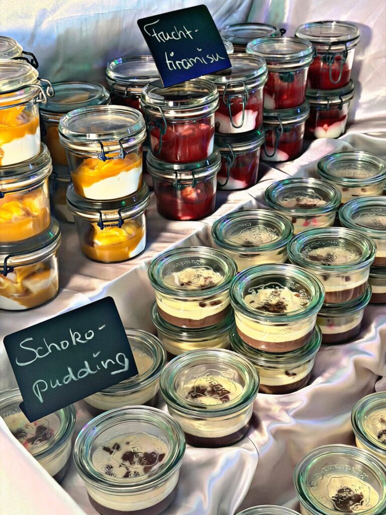 Eine Auslage mit Desserts in Gläsern, darunter Schichten von Tiramisu mit Früchten, gelbes Fruchtparfait und Schokoladenpudding, jeweils mit einem kleinen Kreideschild in deutscher Sprache beschriftet. Die Gläser sind auf einem weißen Tuch angeordnet