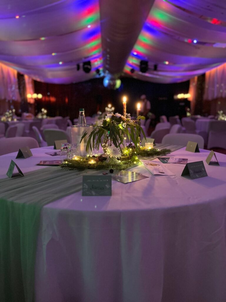 Ein runder Tisch bei einer schwach beleuchteten Veranstaltung ist mit Kerzen, Blumen, einer Glasflasche, Speisekarten und Namenskärtchen dekoriert. Im Hintergrund sind weitere Tische mit ähnlichen Aufbauten und bunten Lichtern an der Decke zu sehen